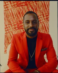 A man wears a red blazer over a black shirt with a red painting behind him.