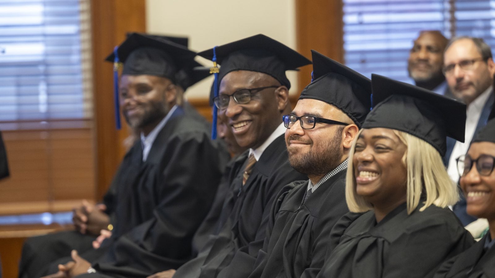 Graduates in cap and gown sit and smile during a graduation ceremony.