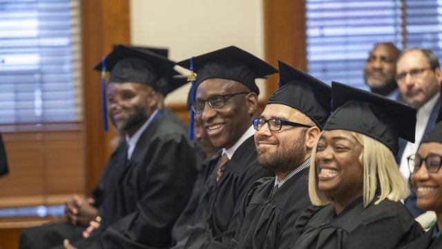 Graduates in cap and gown sit and smile during a graduation ceremony.