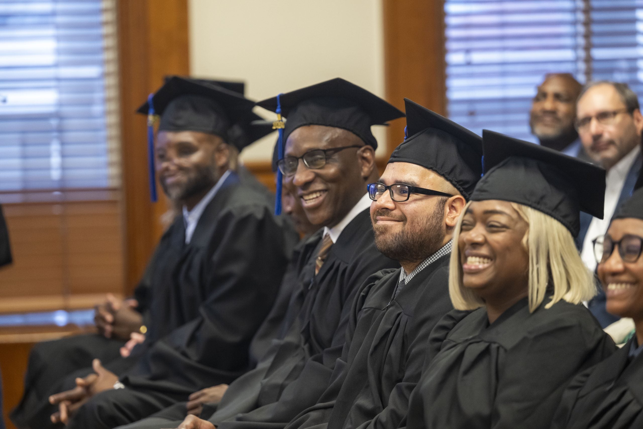 Graduates in cap and gown sit and smile during a graduation ceremony.