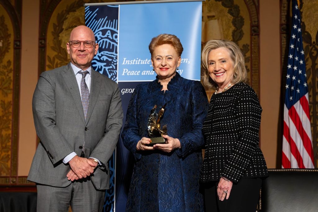 The former president of Lithuania holds an award on a stage. She stands next to Hillary Clinton and Dean Joel Hellman.