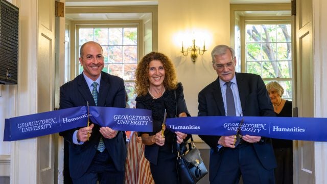 Two men and a woman cut a banner for the Georgetown Humanities Initiative in an academic building.