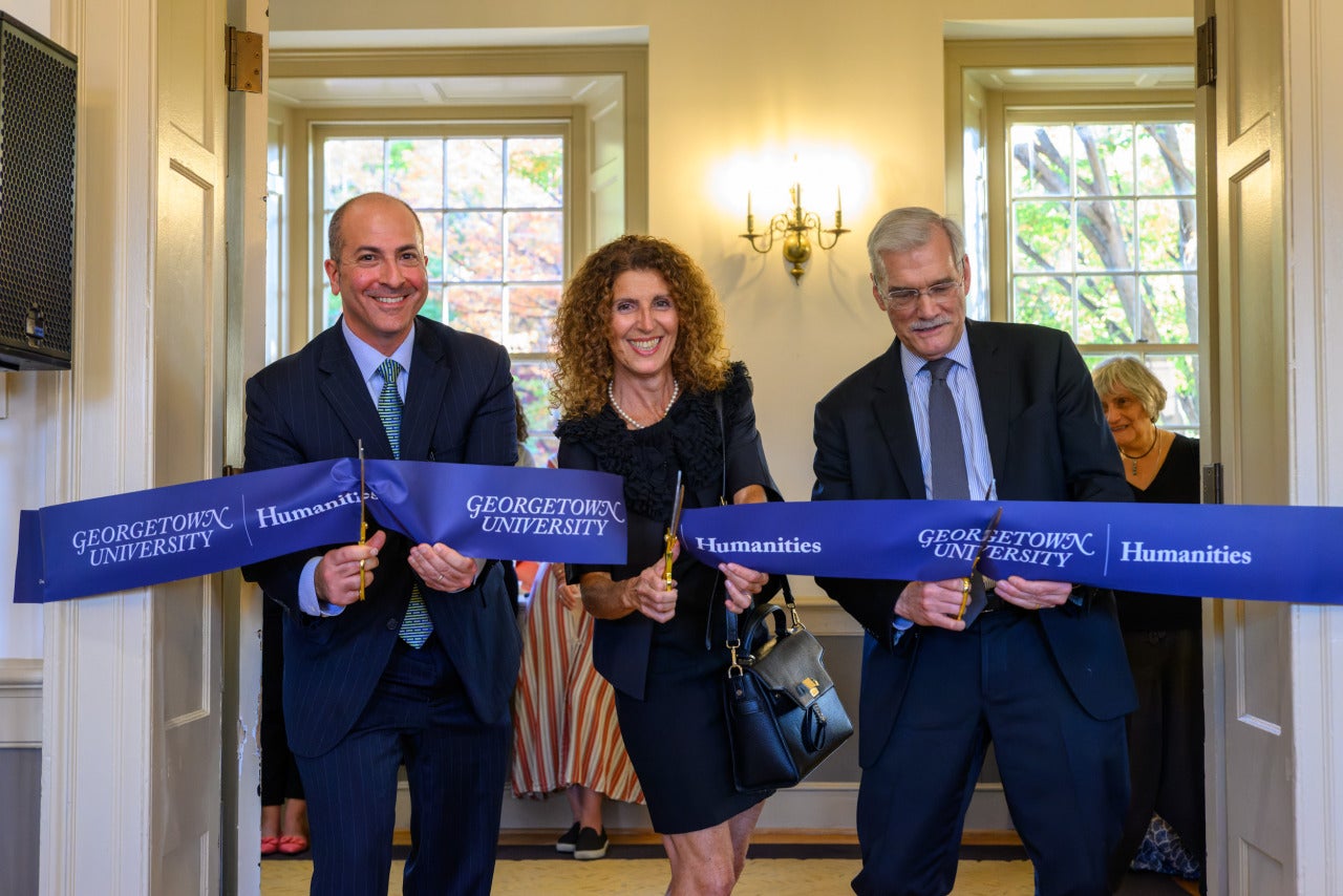 Two men and a woman cut a banner for the Georgetown Humanities Initiative in an academic building.