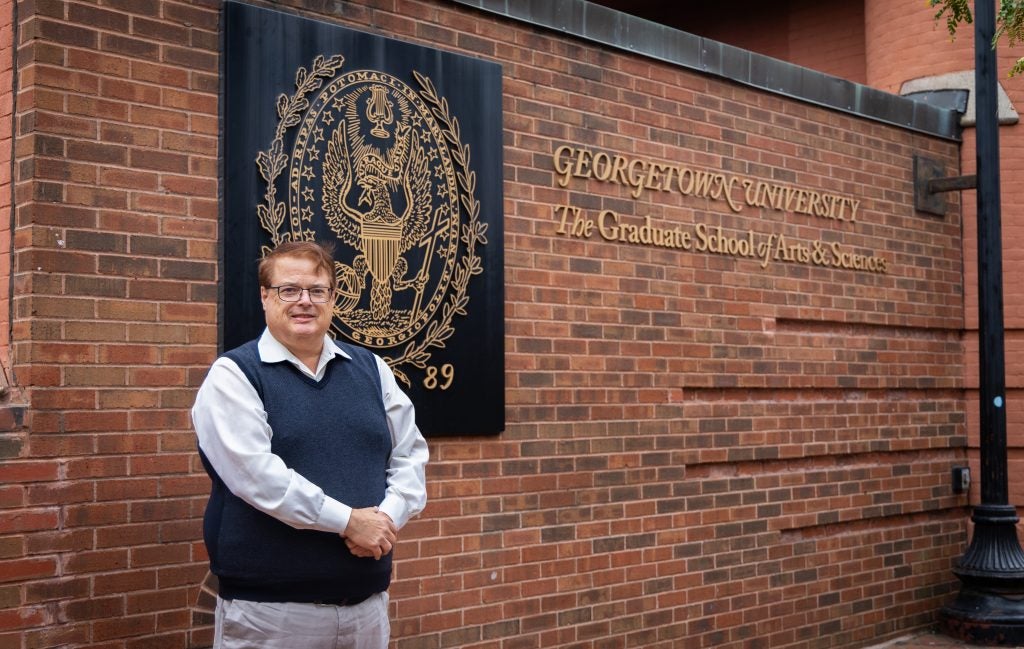 A man with a vest stands with his arms in front of him in front of a brick wall with a seal on it.