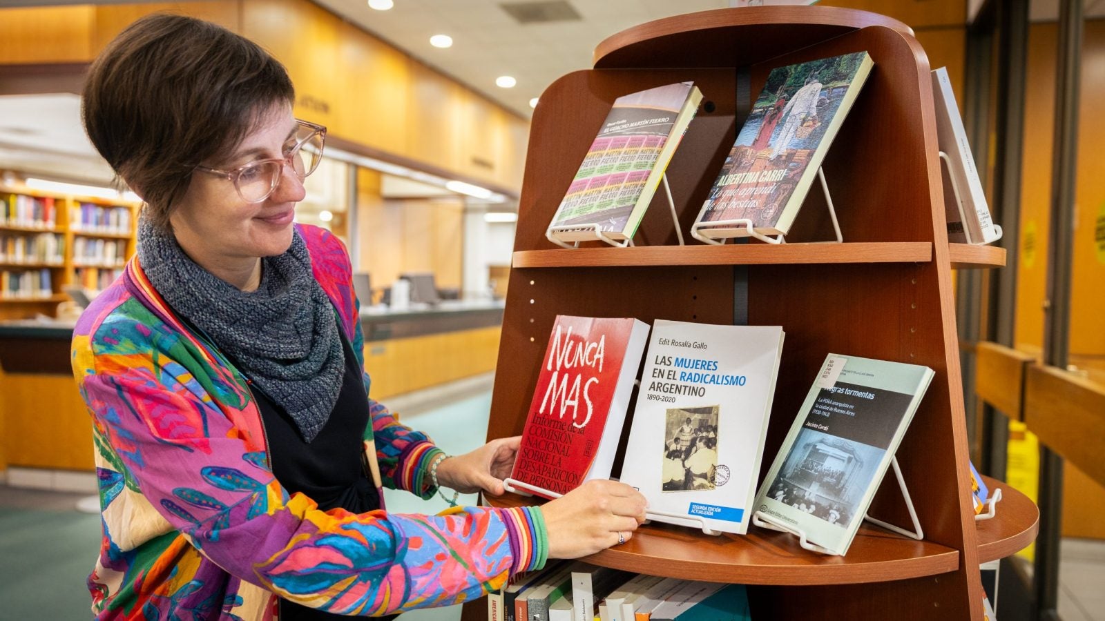 A woman in a colorful pink and blue jacket looks at a book on a shelf in a library.