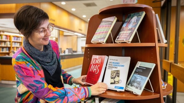 A woman in a colorful pink and blue jacket looks at a book on a shelf in a library.