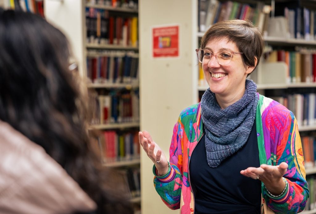 A woman with a pink and blue jacket and a blue scarf smiles with her arms in the air in front of a bookshelf.