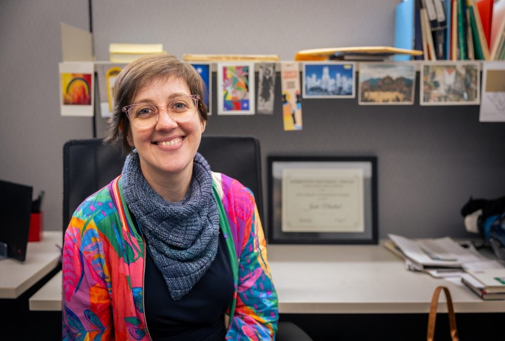 A woman with short brown hair and glasses smiles in a colorful jacket and scarf in front of an office desk.