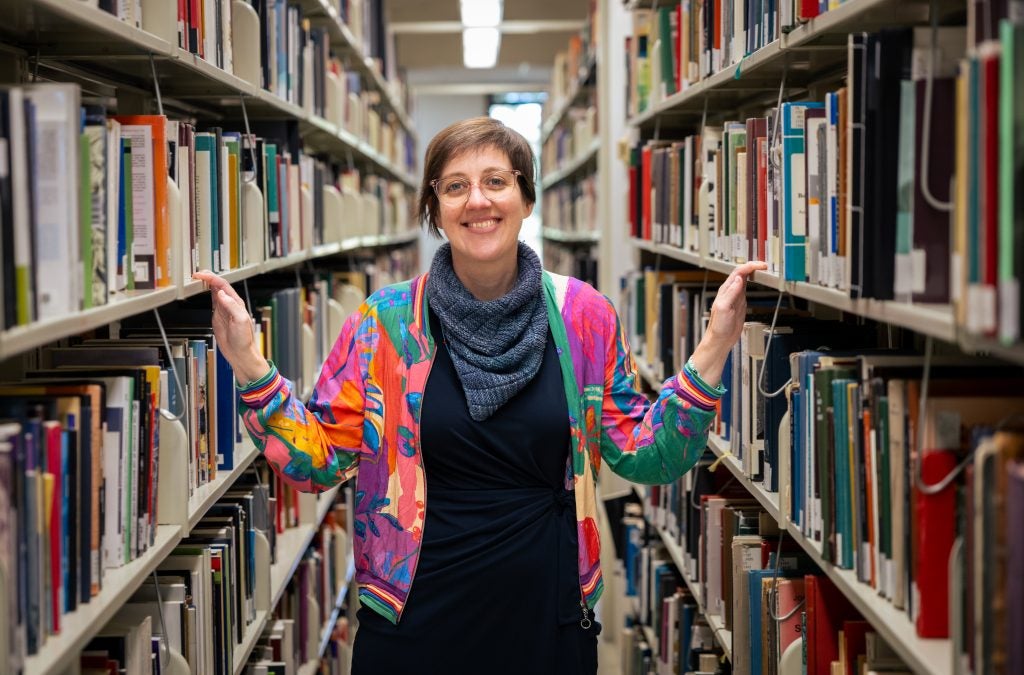 A woman in a pink and blue jacket and a blue scarf stands in between two bookshelves in a library.