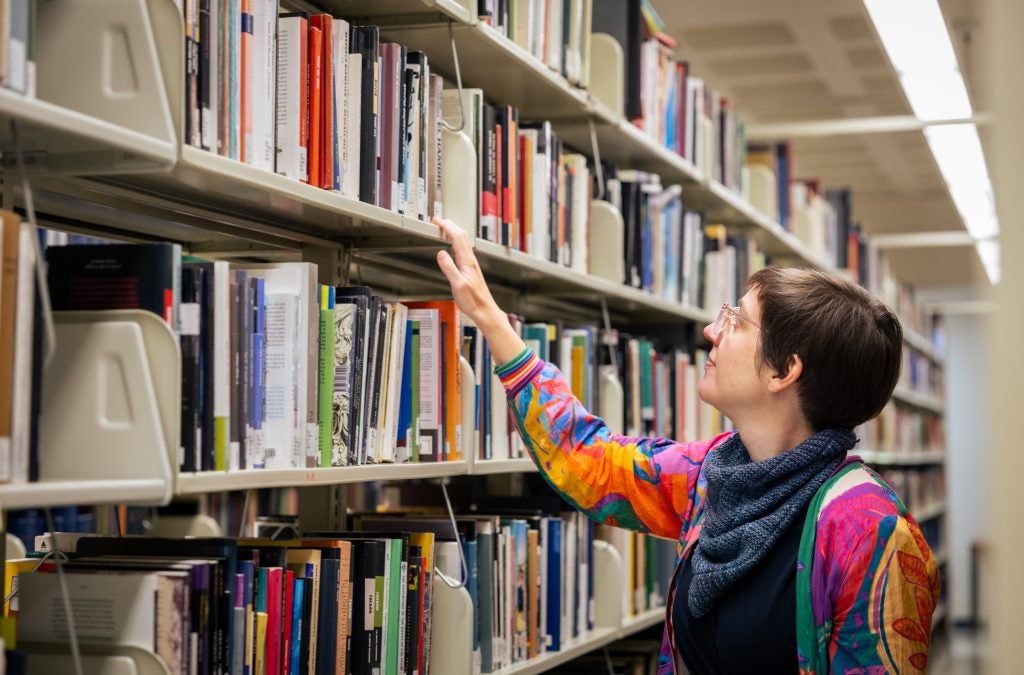 A woman in a colorful jacket and blue scarf looks up at a book shelf in a library.