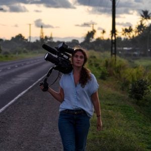 A woman rests a video camera on her shoulder as she walks down a street.