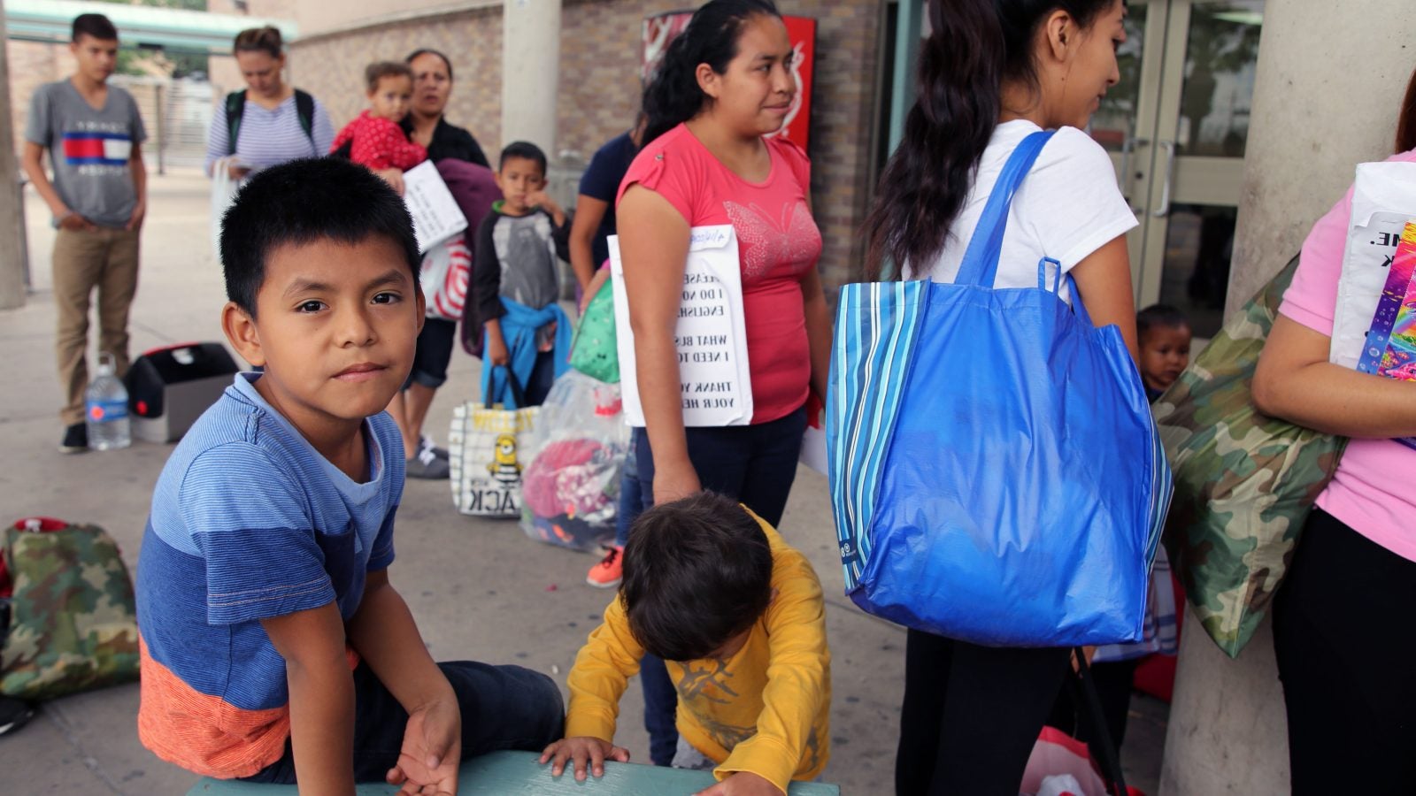 Central American child in blue shirt sits with two women at a port of entry, looking at the camera.