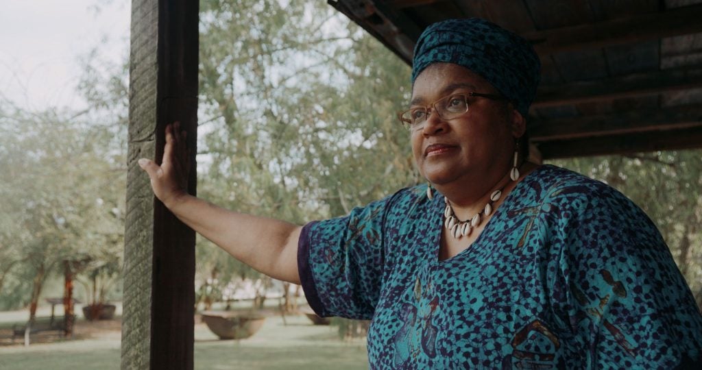 A woman leans against a beam outside a house.