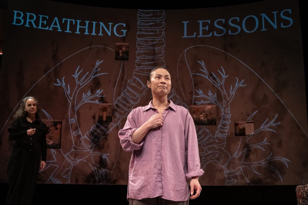 A woman in a purple shirt stands on a stage performing with one fist of her heart. Behind her read the words "Breathing Lessons."