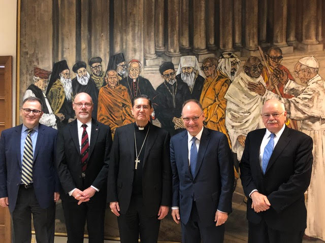 Six mean in formal attire including a Catholic priest in front of a mural