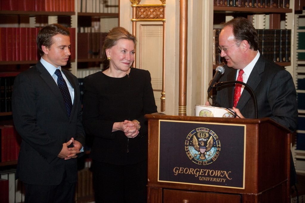 A mother and son stand next to then-president of Georgetown, John J. DeGioia, who's behind a podium in a library.