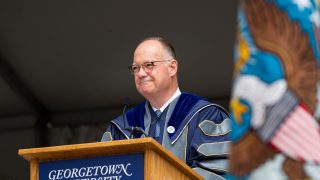 President John DeGioia looks out from a podium that says Georgetown University. He wears academic dress robes.