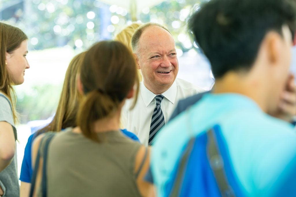 President DeGioia smiles amongst students in a white collared shirt and striped tie.