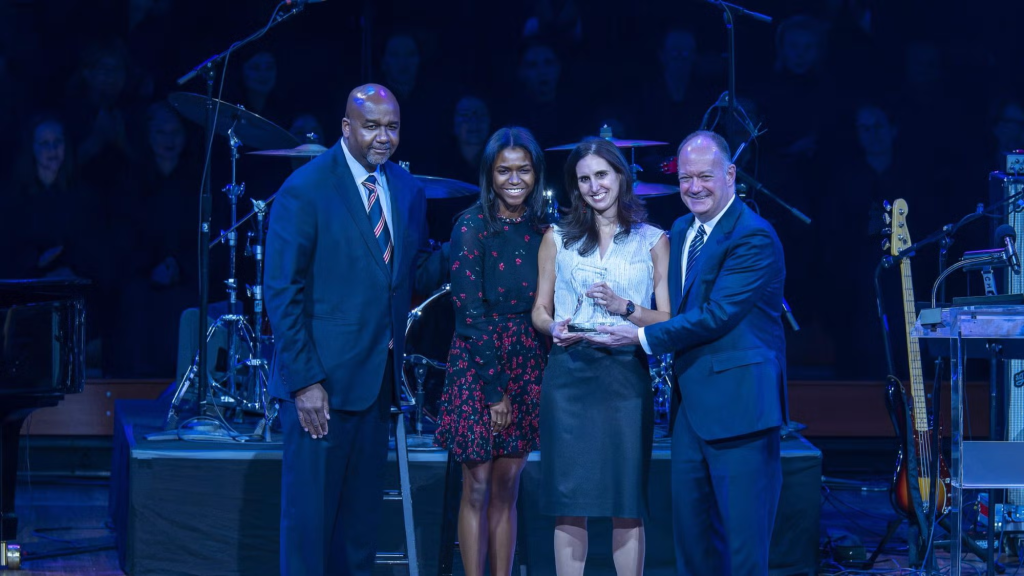 A group of men and women stand onstage of the Kennedy Center in front of a drum set. A woman holds an award in her hands.