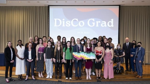 A group of students pose together behind a screen that says "DisCo Grad."