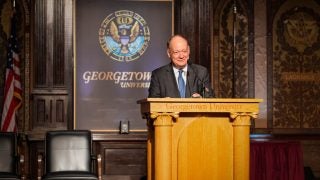 President DeGioia smiles behind a podium in Georgetown&#039;s Gaston Hall.