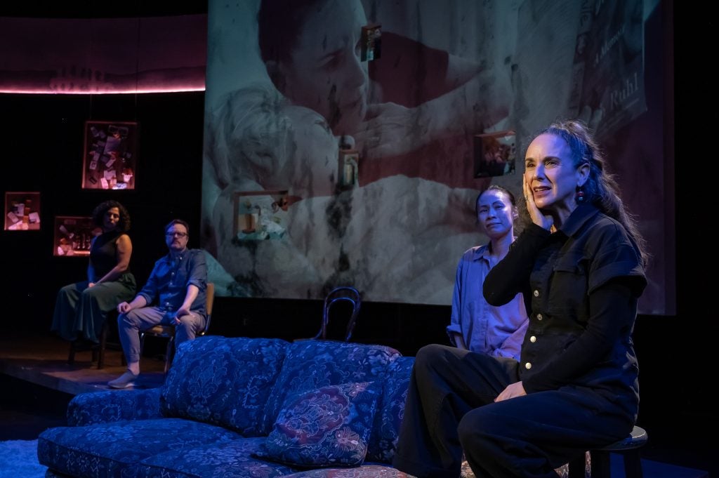 A woman sits on a sofa and cups one hand over her cheek as she performs in a play.