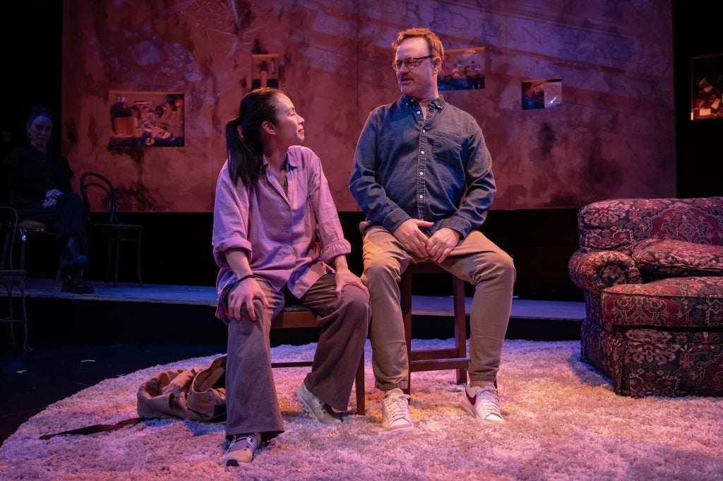 A man and a woman sit on chairs next to each other on a stage while performing a play.