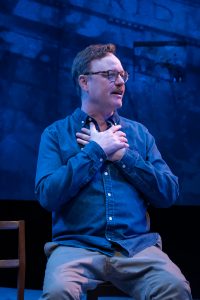 A man wearing glasses and a button-down shirt crosses his hands over his heart while seated on a stage.