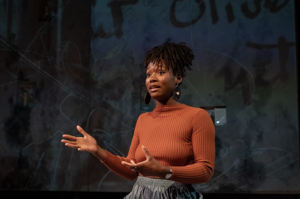 A woman stands on a stage with her arms slightly outstretched while performing a play.