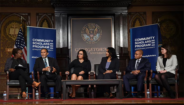 A group of panelists sit on a stage and discuss. Behind them are signs that say "Community Scholars Program."