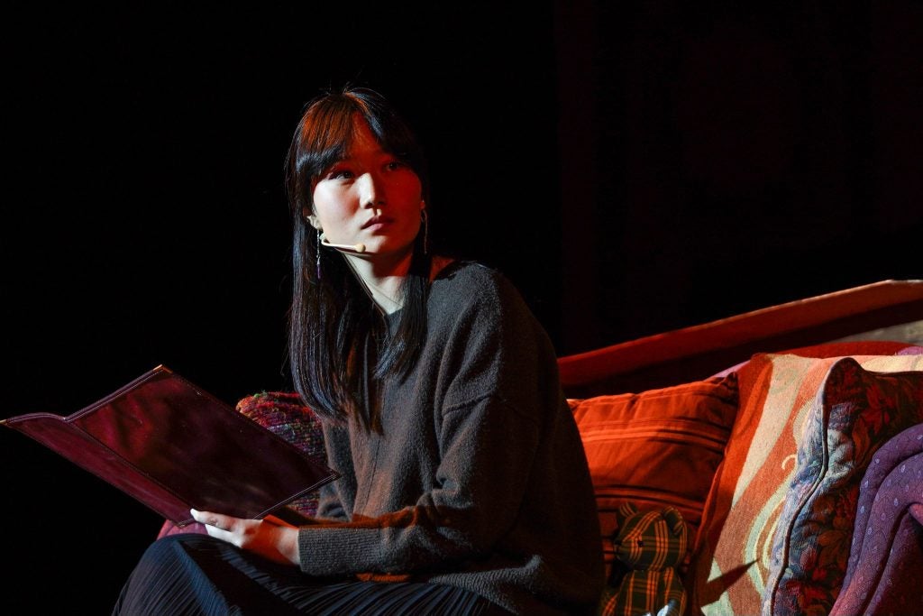 A woman with a microphone sits on a stage while performing in a role. She holds a book.