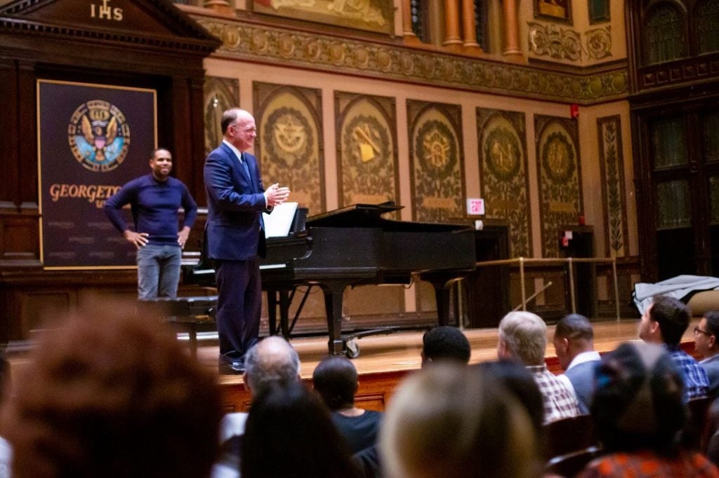 President John DeGioia stands on a stage in Gaston Hall in front of a piano. He speaks to an audience in front of him.