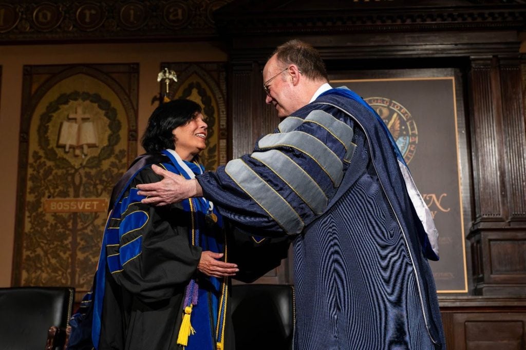 President DeGioia, dressed in a blue and gray academic robe, greets a professor in a black academic robe.