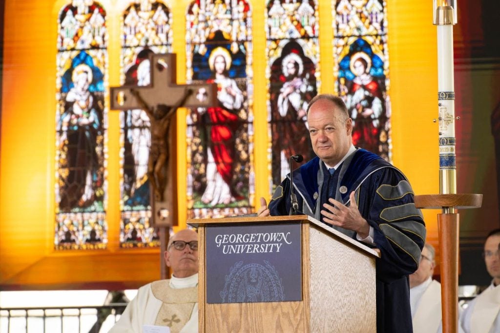 John DeGioia stands in front of a pulpit that says "Georgetown University" in front of stained glass windows in chapel.