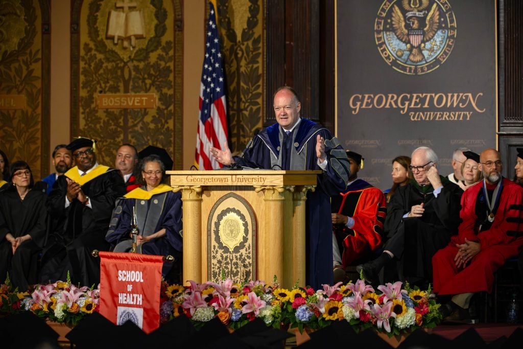 President DeGioia speaks behind a podium in an academic robe while other faculty members in academic robes listen on the stage.