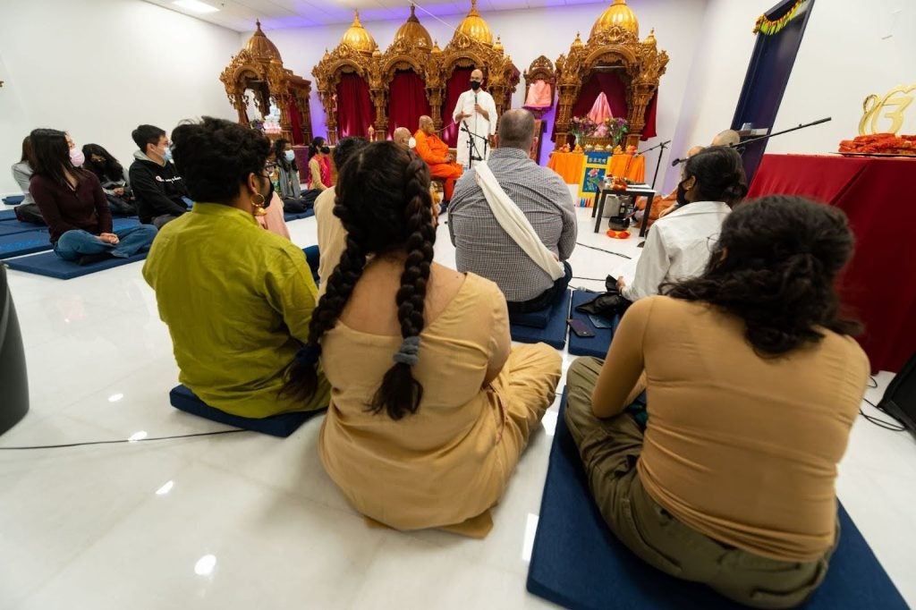 A group of students sit on the floor inside a white room with a teak shrine at the front of the room.