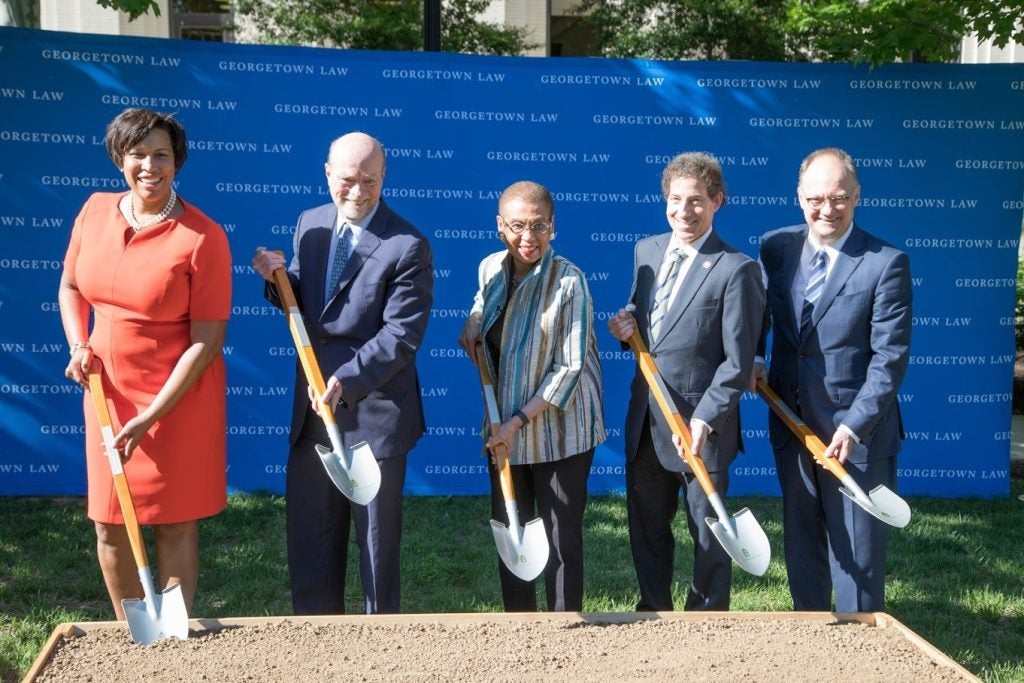 Two women and three men hold shovels and pose in front of a blue banner that says "Georgetown Law."