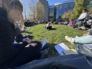 A circle of students sit outside of Hariri with their laptops on a sunny day