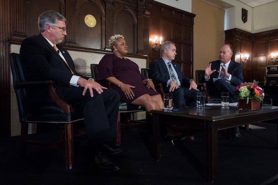 A group of presidents of local community colleges and universities sit together on a stage listening and talking.