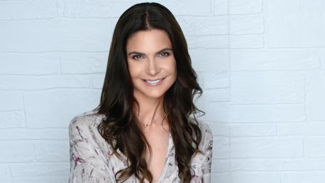 A woman with brown hair smiles against a white brick background.