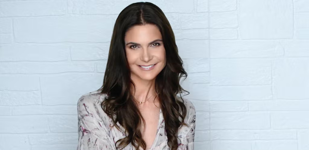 A woman with brown hair smiles against a white brick background.