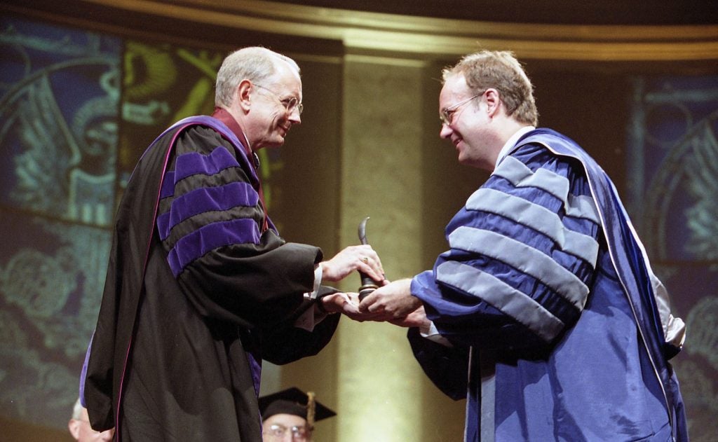 A man in a purple and black academic robe presents a gift to another man in a striped purple and gray academic robe.