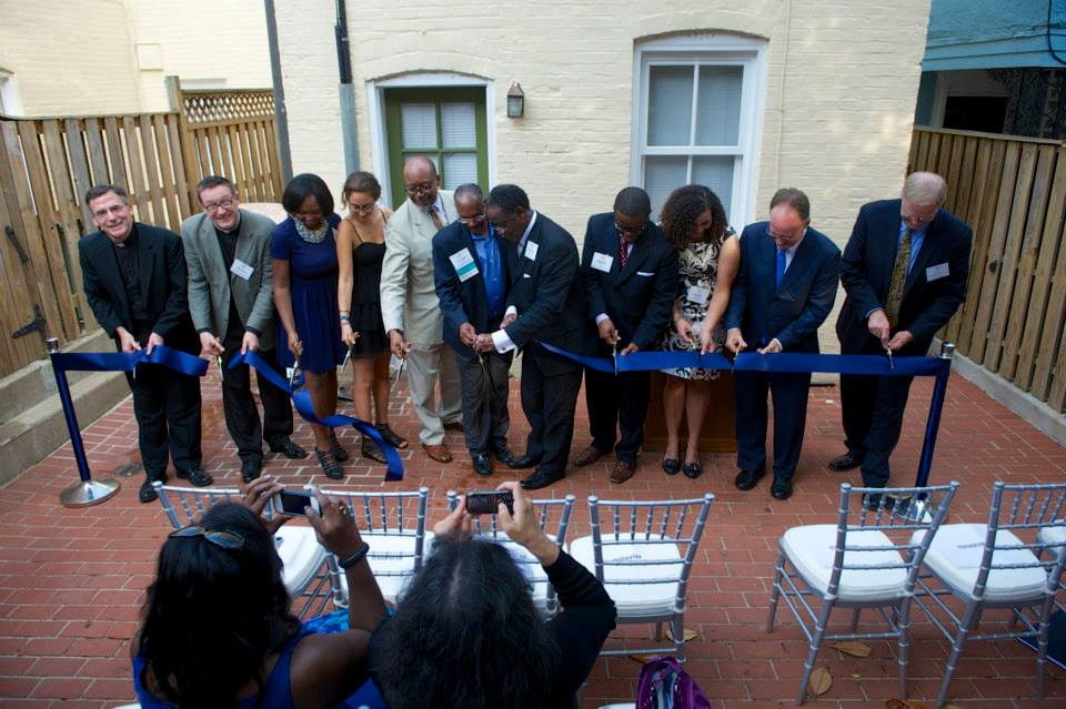 A group of alumni and community members cut a blue ribbon on a patio.