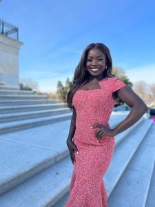 A Black woman smiles in a pink dress on steps in Washington, DC.