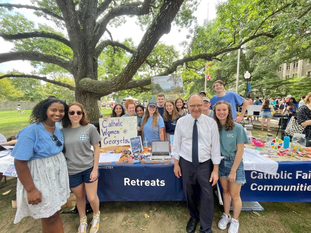 John DeGioia stands with students in front of a campus ministry booth.