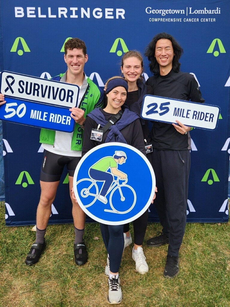 Two women and two men pose in front of a blue banner holding signs like "Cancer Survivor: 50 Mile Rider" and "25 Mile Rider"