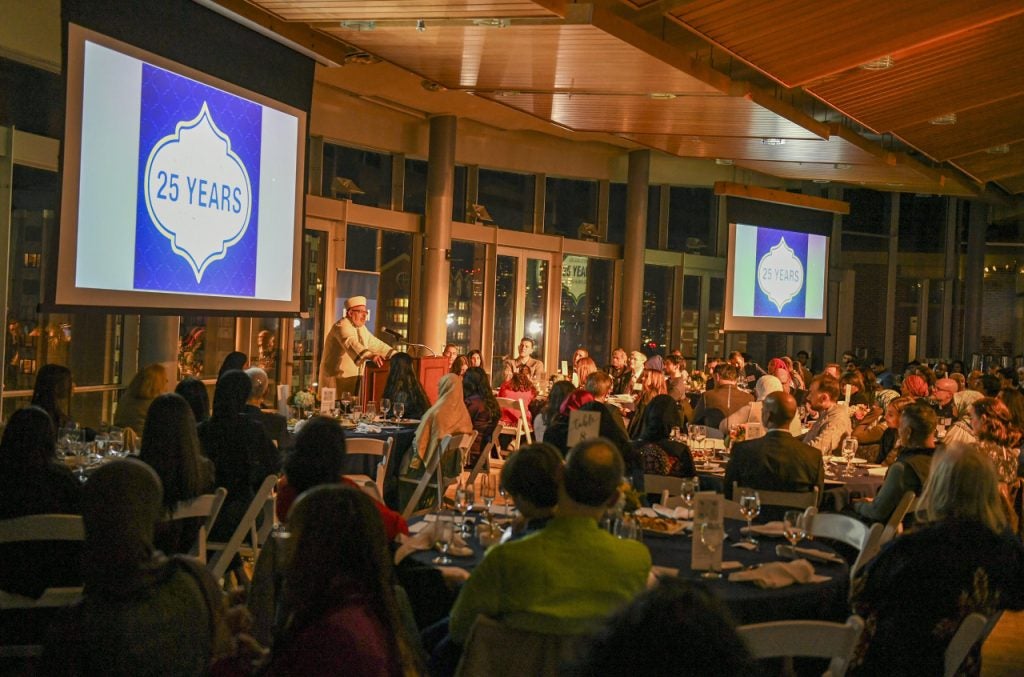 A crowd seated around circular tables listen to Imam Yahya Hendi, who stands behind a podium and delivers remarks at the 25th anniversary of Muslim Life.