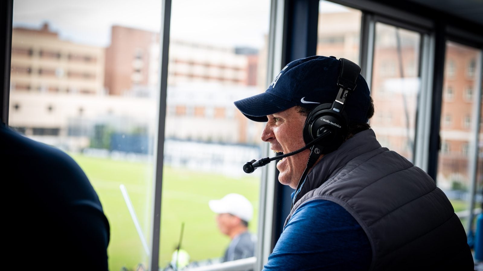 Dan Helfrich smiles and cheers looking out from a press box onto a soccer field.