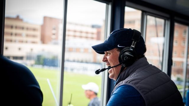 Dan Helfrich smiles and cheers looking out from a press box onto a soccer field.