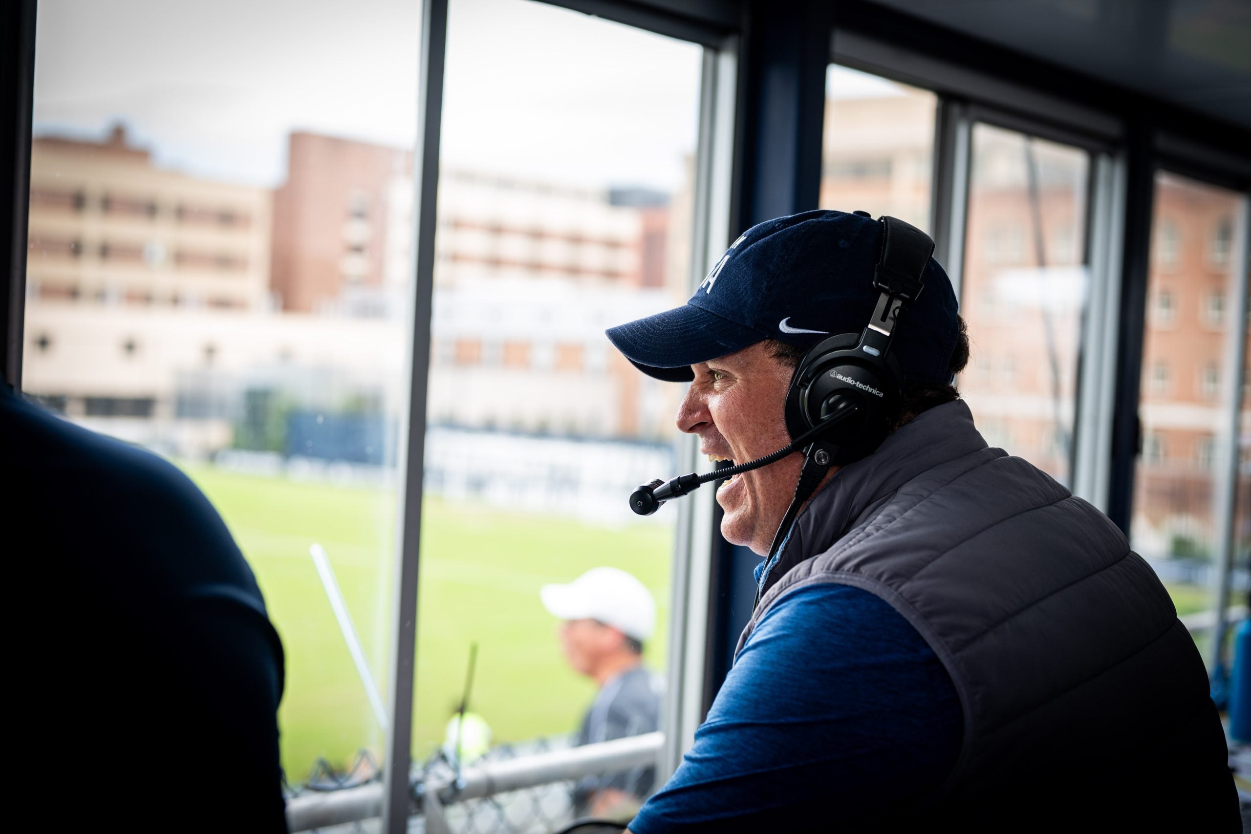 Dan Helfrich smiles and cheers looking out from a press box onto a soccer field.